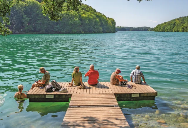 Personen sitzen in der Sonne auf einem Steg am Wasser