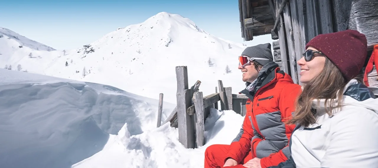 Zwei Personen neben einem Holzgebäude vor verschneitem Berg in Ski amadé.