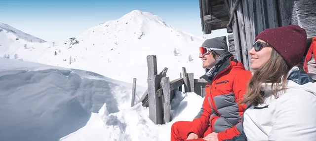 Zwei Personen neben einem Holzgebäude vor verschneitem Berg in Ski amadé.