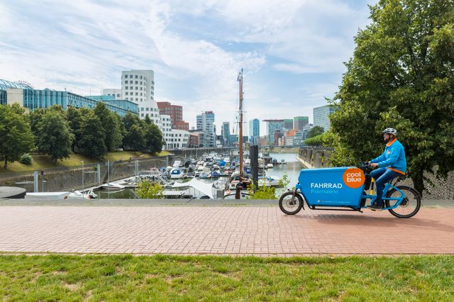 Coolblue-Fahrer auf einem blauen Cargo-Bike am Hafen in Düsseldorf, im Hintergrund Boote, Bäume und Bürogebäude.