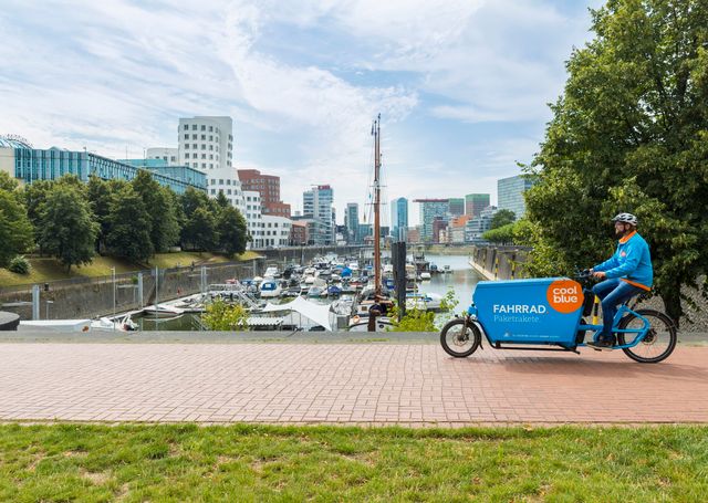 Coolblue-Fahrer auf einem blauen Cargo-Bike am Hafen in Düsseldorf, im Hintergrund Boote, Bäume und Bürogebäude.