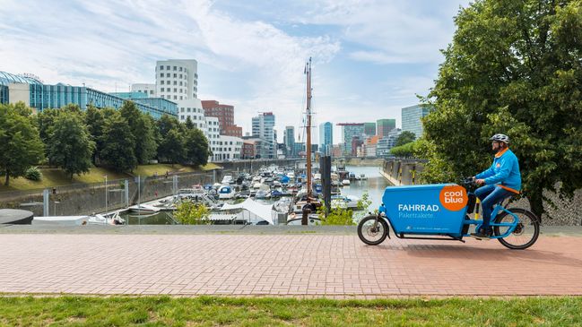 Coolblue-Fahrer auf einem blauen Cargo-Bike am Hafen in Düsseldorf, im Hintergrund Boote, Bäume und Bürogebäude.