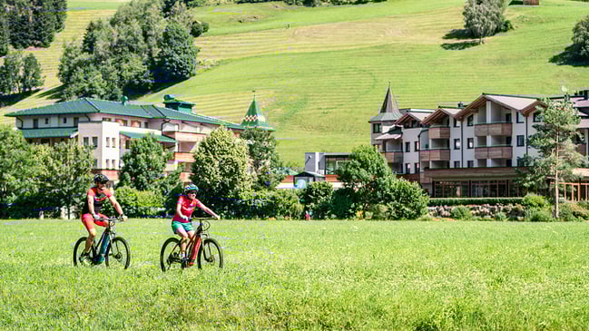 Zwei Fahrradfahrende in der grünen Natur mit Gebäuden im Hintergrund.