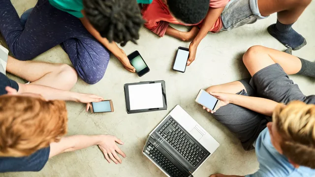 Gruppe von Kindern sitzt im Kreis auf dem Boden und schaut auf Smartphones, ein Tablet und einen Laptop in der Mitte.